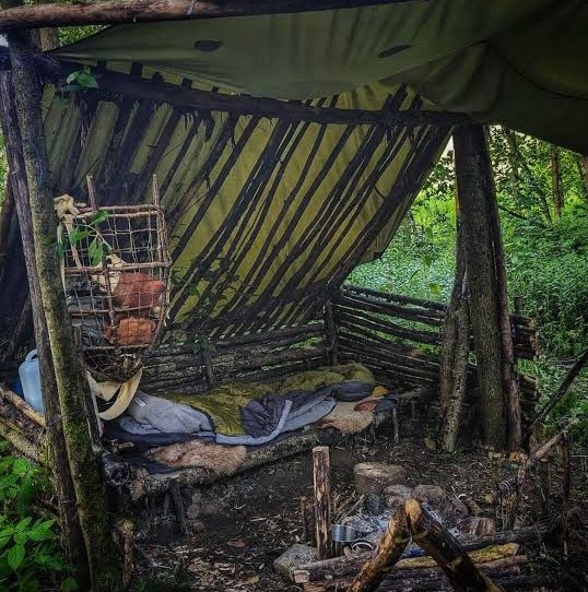 Lived-in bushcraft camp under a green tarp with a raised pole bed, sleeping bag, hide and a woven basket