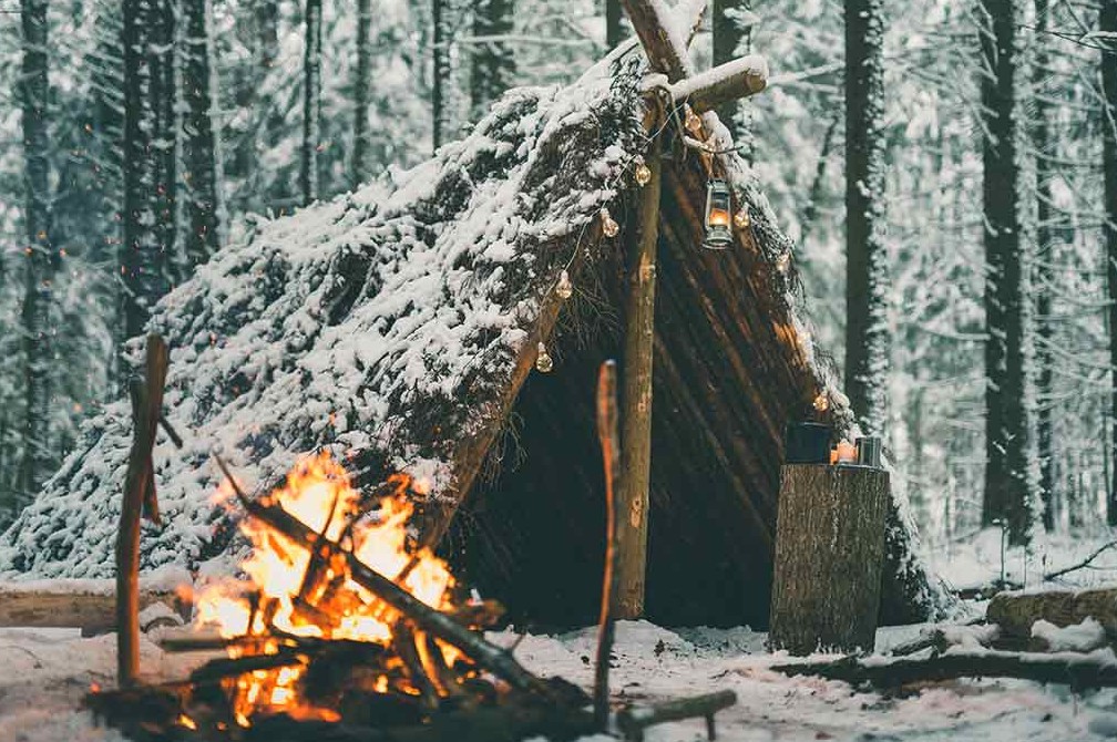 Snow covered lean-to shelter in a winter pine forest with a fire burning in front and a lantern hung from the ridge pole