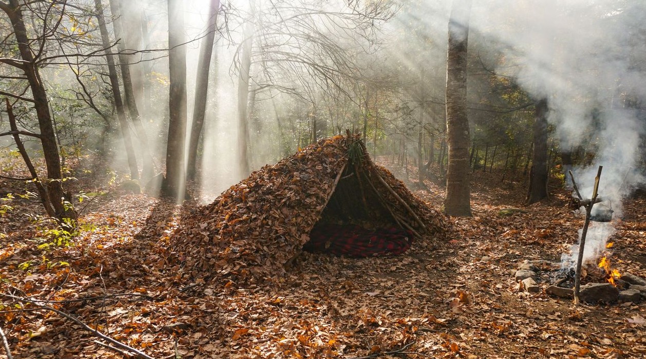 Debris hut piled deep with oak leaves in an autumn hardwood forest, sunbeams and campfire smoke behind it