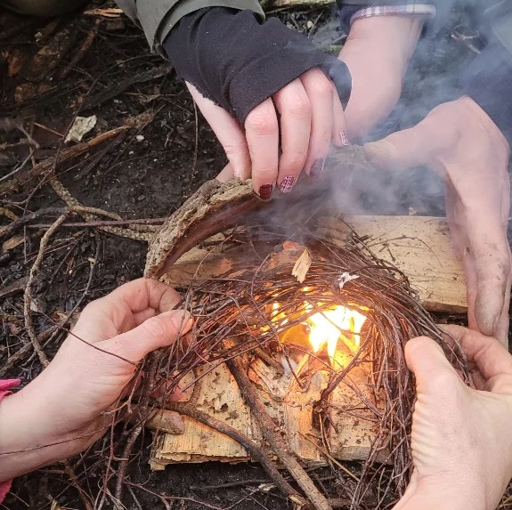Two pairs of hands feeding birch bark into a smoking tinder bundle as the first flame catches