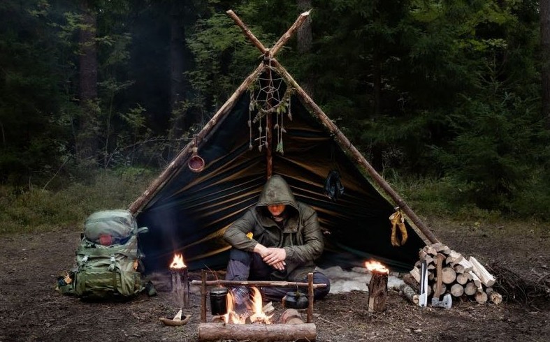 Camper in a hooded jacket sitting under a dark tarp lean-to beside a campfire, with split firewood stacked and an axe nearby