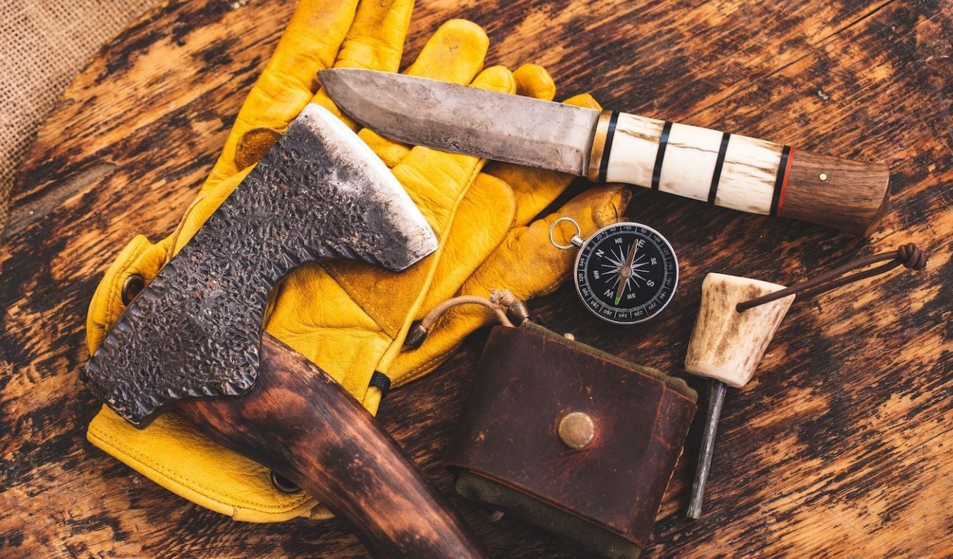 Forged hatchet and a hunting knife with a stacked bone handle on weathered wood, with leather gloves, a compass and an antler handled awl
